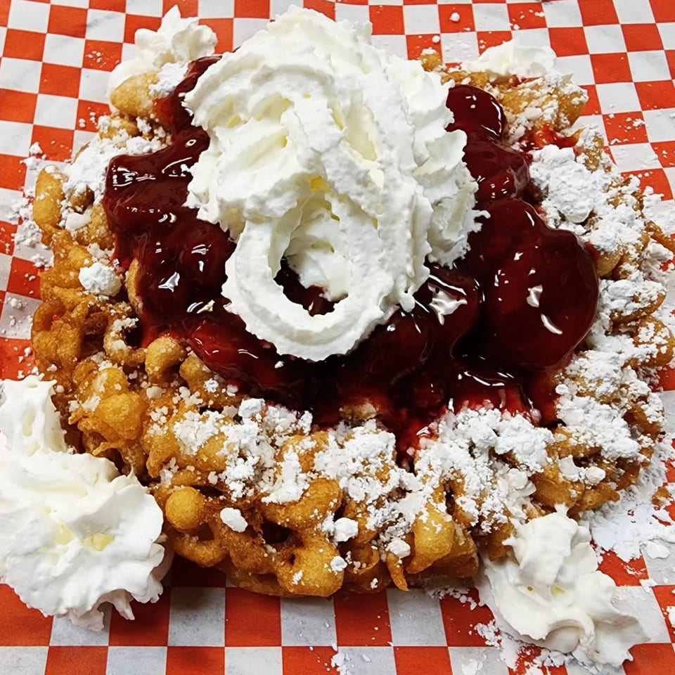 Funnel cake topped with cherry pie filling, whipped cream, and powdered sugar on a checkered paper.