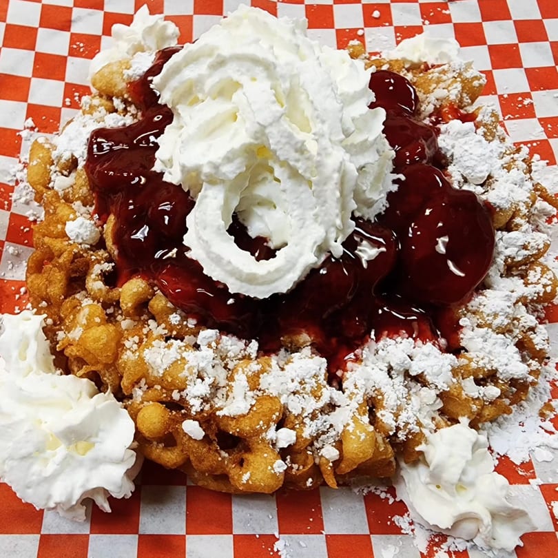 Funnel cake topped with cherry pie filling, whipped cream, and powdered sugar on a checkered paper.