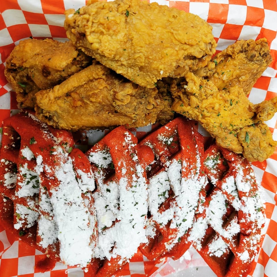 Golden fried chicken pieces and red velvet waffles with powdered sugar on red and white checkered paper.