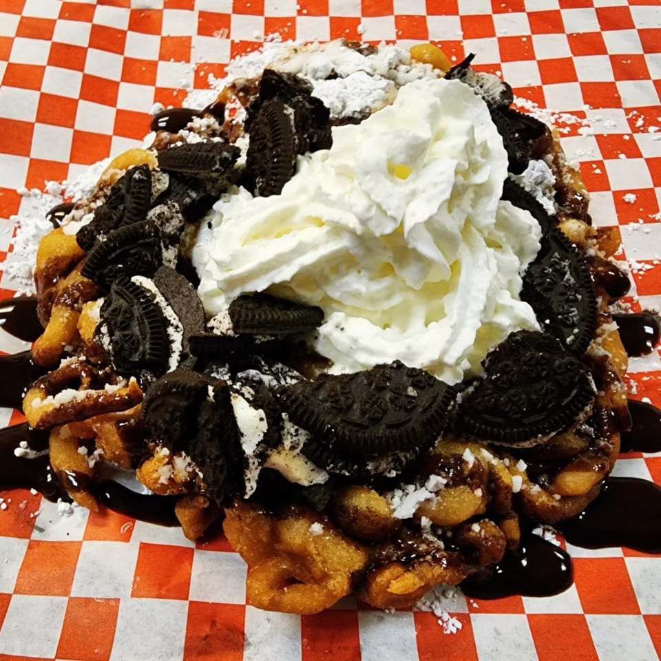Funnel cake topped with whipped cream, Oreo cookies, chocolate syrup, and powdered sugar on checkered paper.