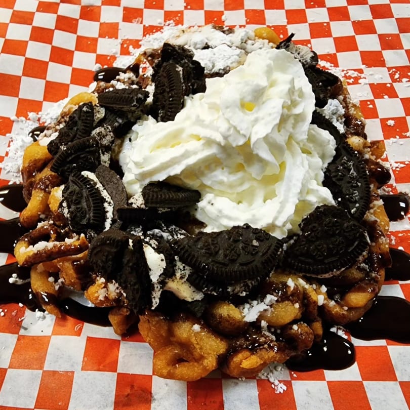 Funnel cake topped with whipped cream, Oreo cookies, chocolate syrup, and powdered sugar on checkered paper.
