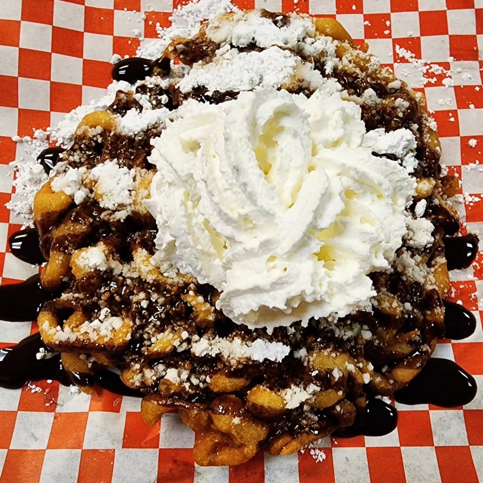Funnel cake topped with chocolate syrup, powdered sugar, and whipped cream on a red and white checkered paper.