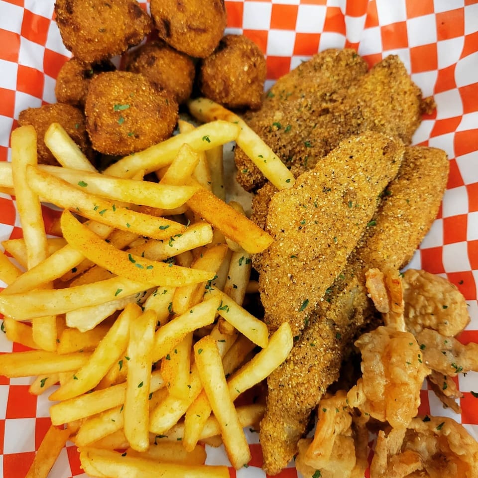 A basket of Louisiana Famous Fried Chicken with fries, hush puppies, and fried fish on red and white checkered paper.